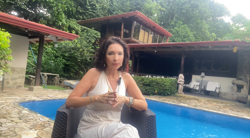 <img data-lazyloaded= Claudia Morassutti hablando sobre la proyección de la sombra en la relación de pareja, sentada junto a una piscina con un entorno natural de fondo.