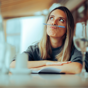 Mujer sentada en una cafetería con los brazos cruzados sobre la mesa y un cuaderno abierto, transmitiendo un momento de reflexión y pausa consciente.